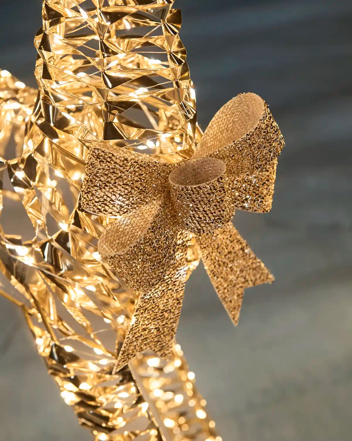 A close-up of a gold decorative glitter bow on LED reindeers neck along with warm white LED lights against snowy grass