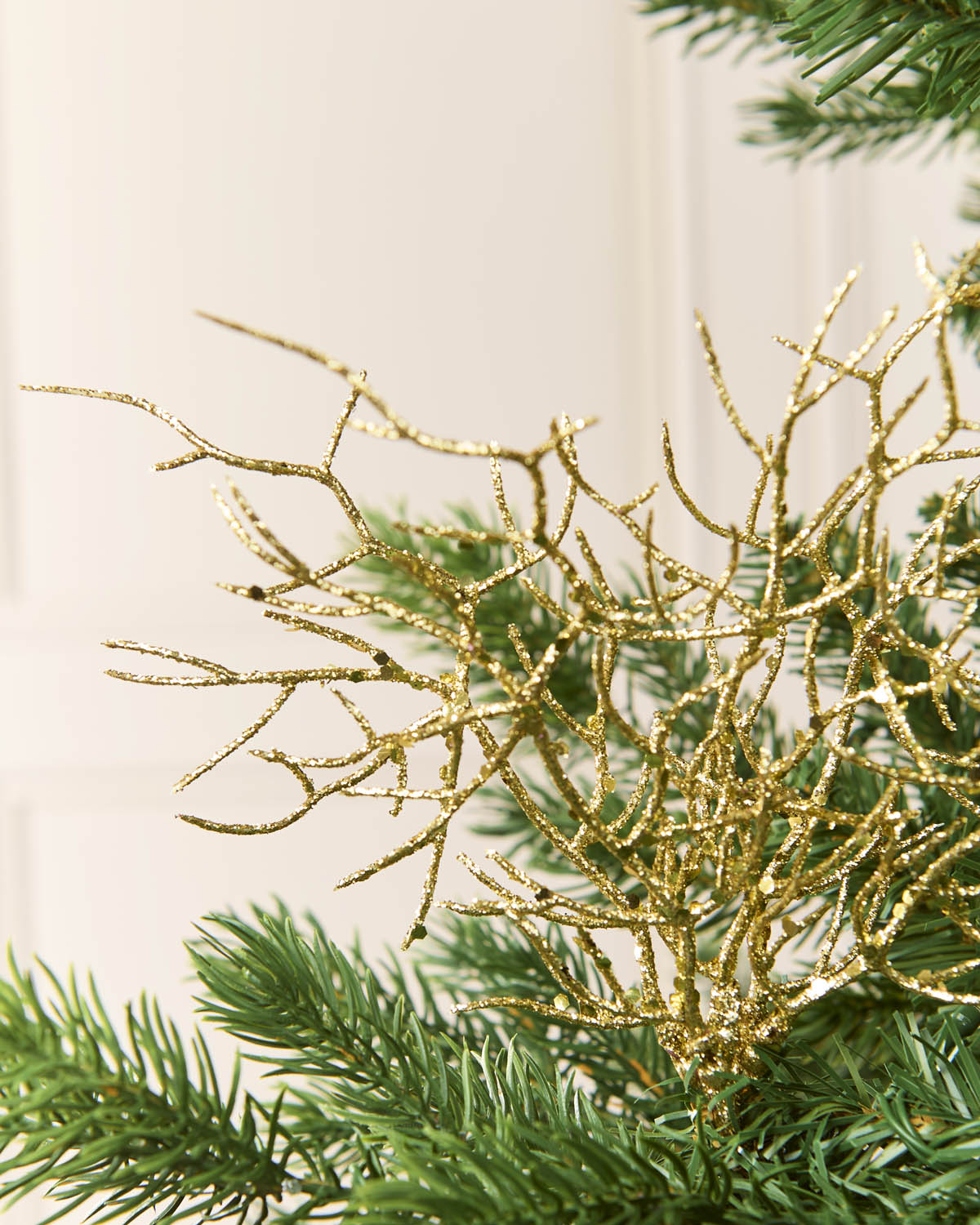 A close-up of a gold glittery branch or coral-style decorative ornament layered among green pine needles against a soft cream background.