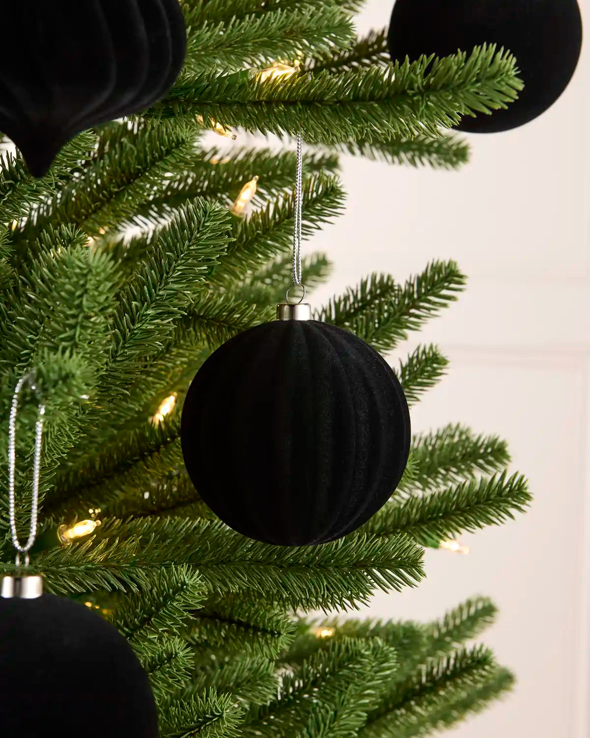 A close-up of a green Christmas tree decorated with black ribbed velvet ball ornaments and warm white string lights against a light neutral background.