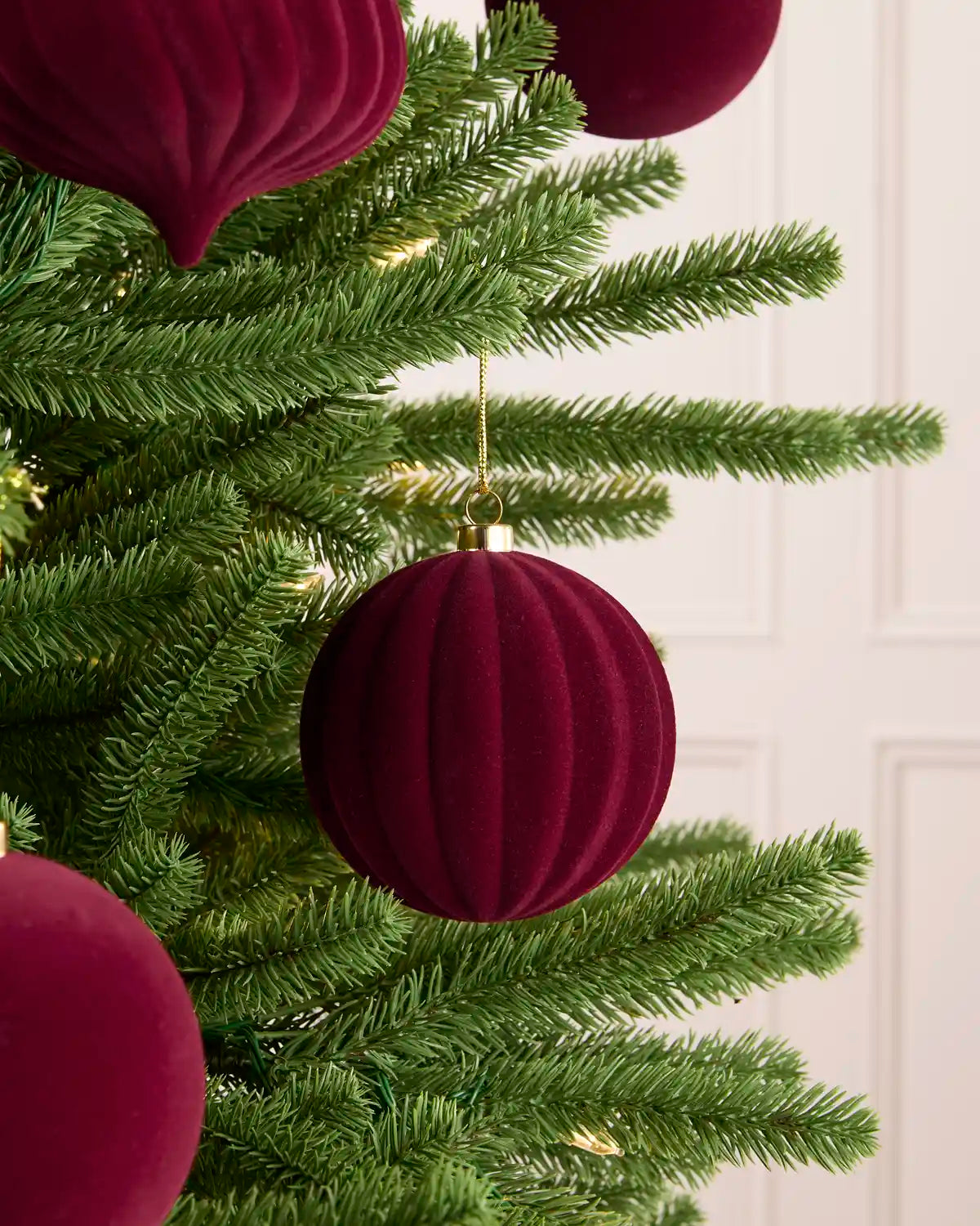 A close-up of a green Christmas tree decorated with burgundy ribbed velvet ball ornaments and matching hanging ornaments, with soft warm lights visible in the branches.