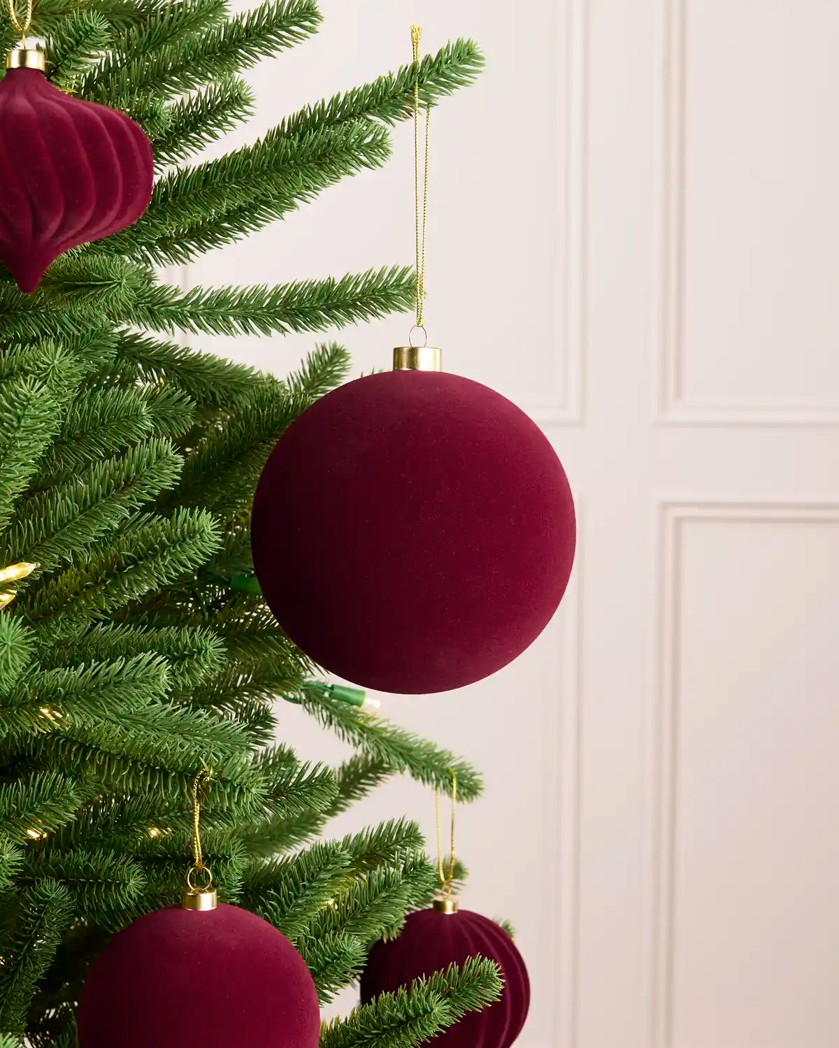 A close-up of a green Christmas tree decorated with deep burgundy velvet ball and ribbed ball ornaments, hanging against a light cream paneled wall.