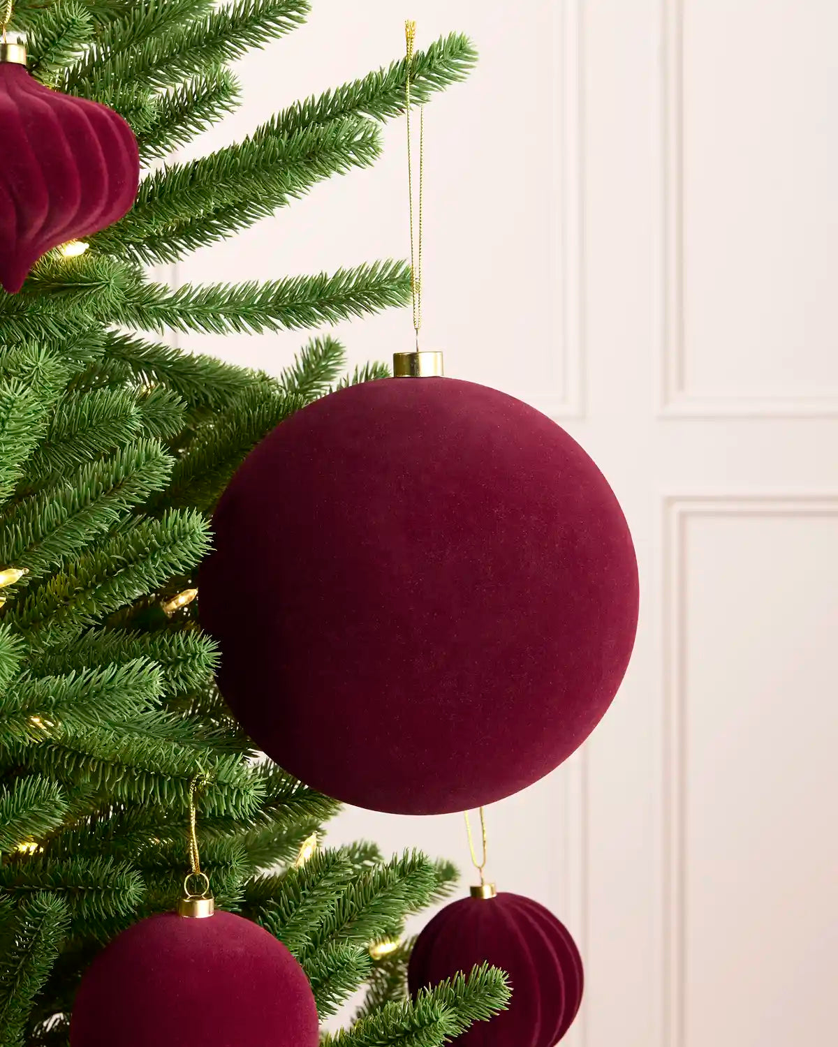 A close-up of a green Christmas tree decorated with large, deep burgundy velvet spherical ornaments and smaller ribbed ornaments, with a soft cream paneled wall in the background.