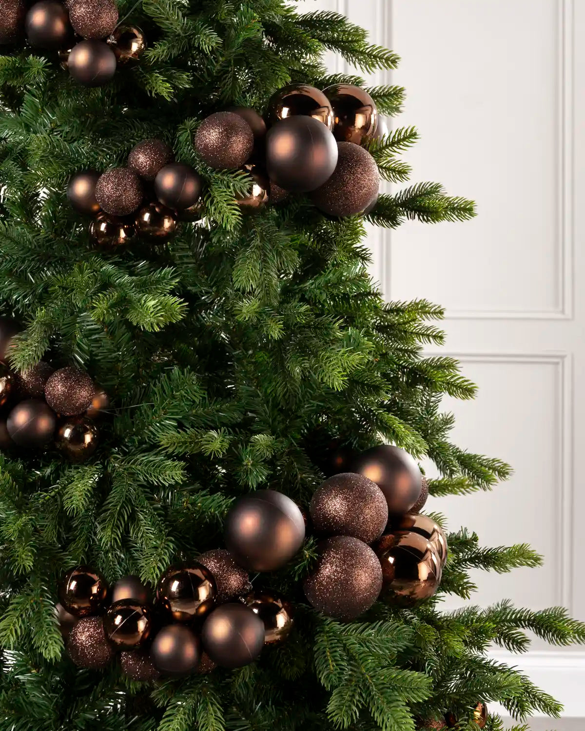 A close-up of a green Christmas tree decorated with metallic Espresso coloured spherical ornaments, some with a smooth matte finish and others with a glittery texture, set against a white paneled wall.