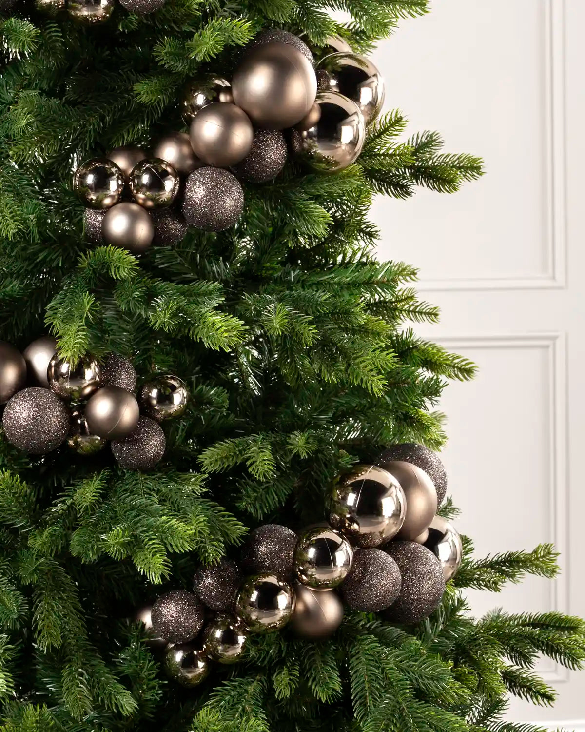 A close-up of a green Christmas tree decorated with metallic and glittery spherical ornaments in mocha set against a white paneled wall.