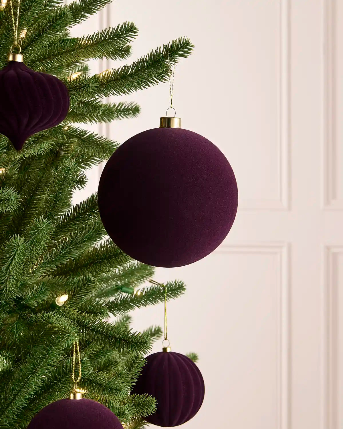 A close-up of a green Christmas tree decorated with purple velvet spherical ornaments and one teardrop-shaped ornament, hanging against a light cream panelled wall.