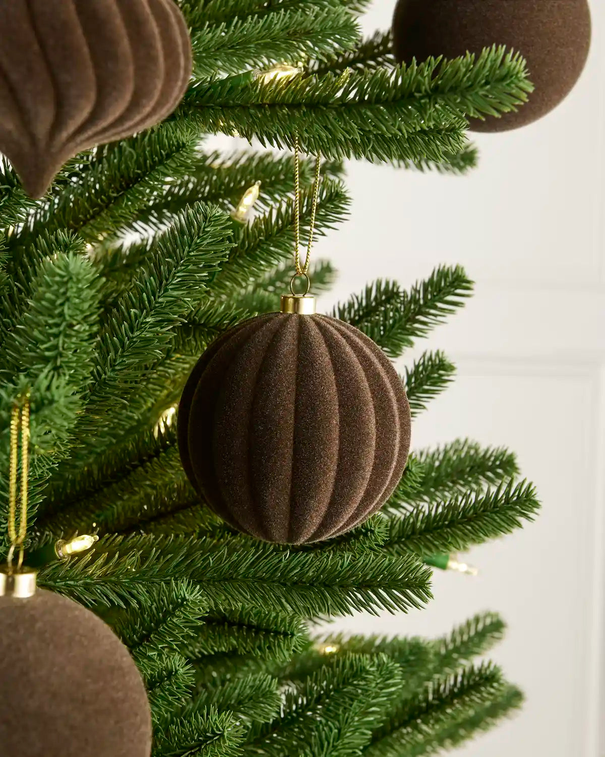 A close-up of a green Christmas tree decorated with ribbed brown felt ornaments and small warm white fairy lights against a light neutral background.