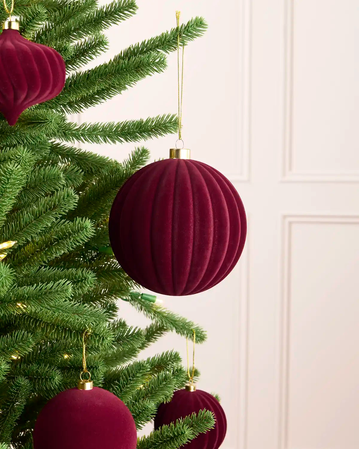 A close-up of a green Christmas tree decorated with ribbed deep burgundy velvet ball ornaments and gold hanging loops against a soft cream paneled background.
