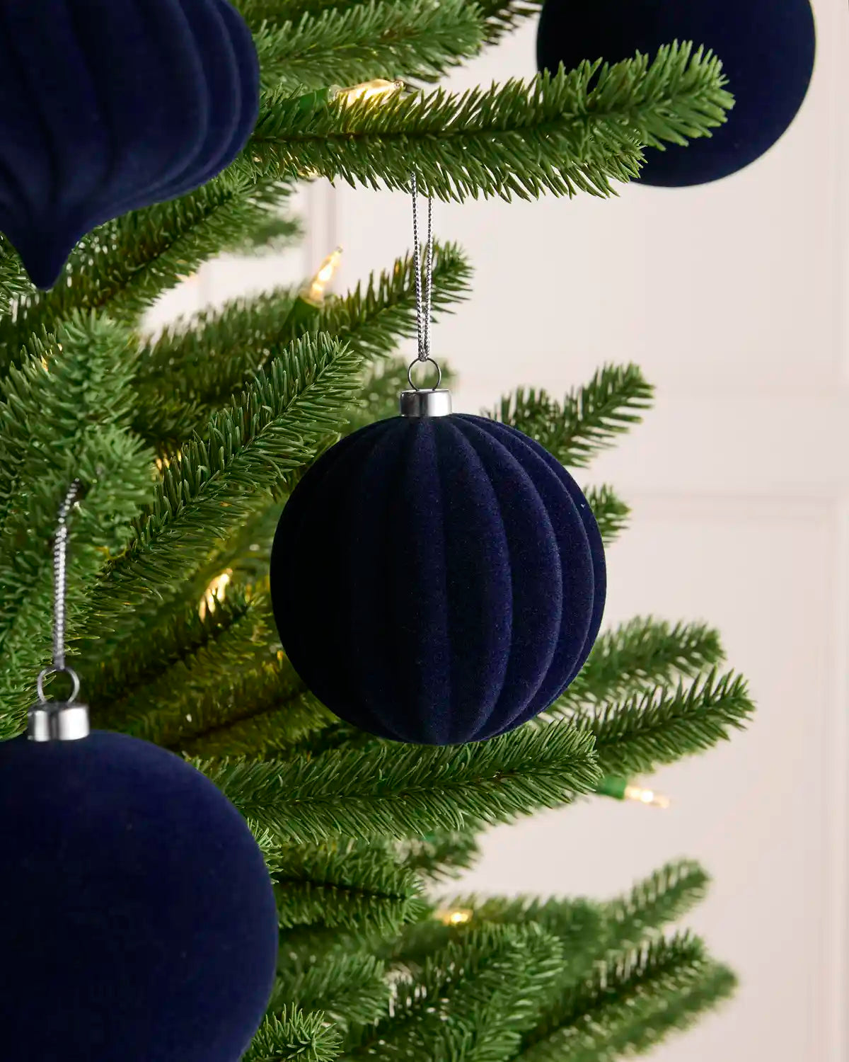 A close-up of a green Christmas tree decorated with ribbed navy blue velvet ball ornaments and small warm white lights against a light neutral background.