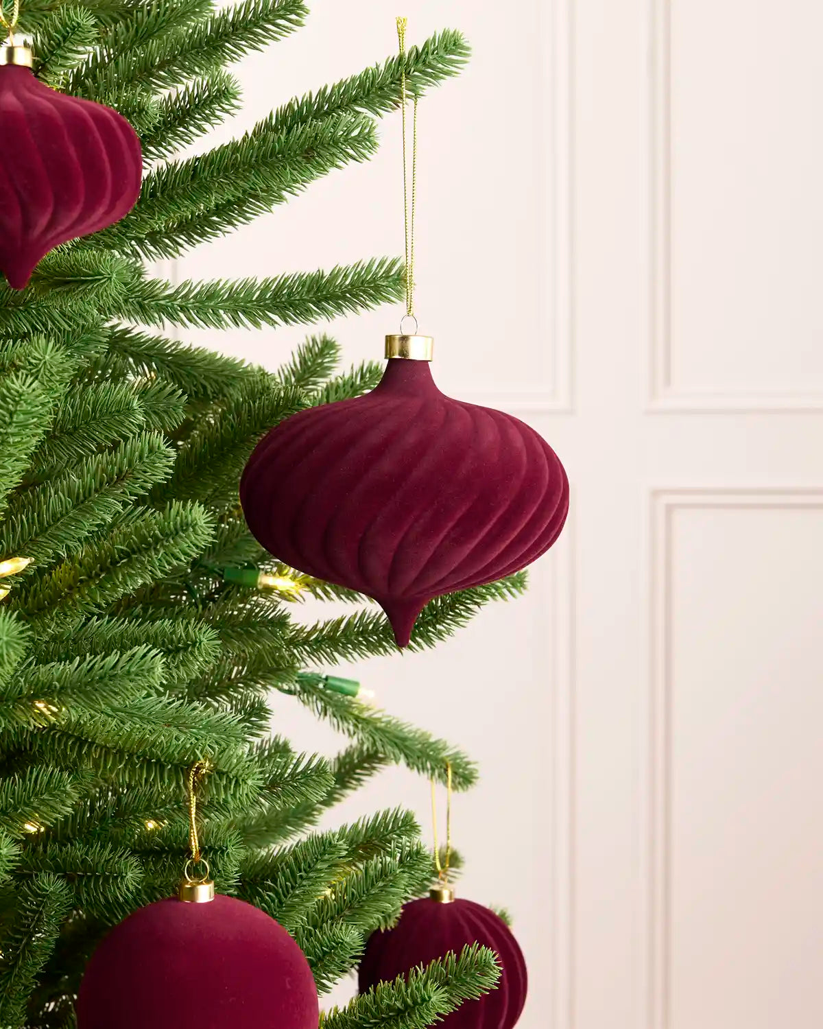 A close-up of a green Christmas tree decorated with ribbed, onion-shaped burgundy velvet ornaments with gold caps and gold hanging strings, set against a light cream paneled wall.