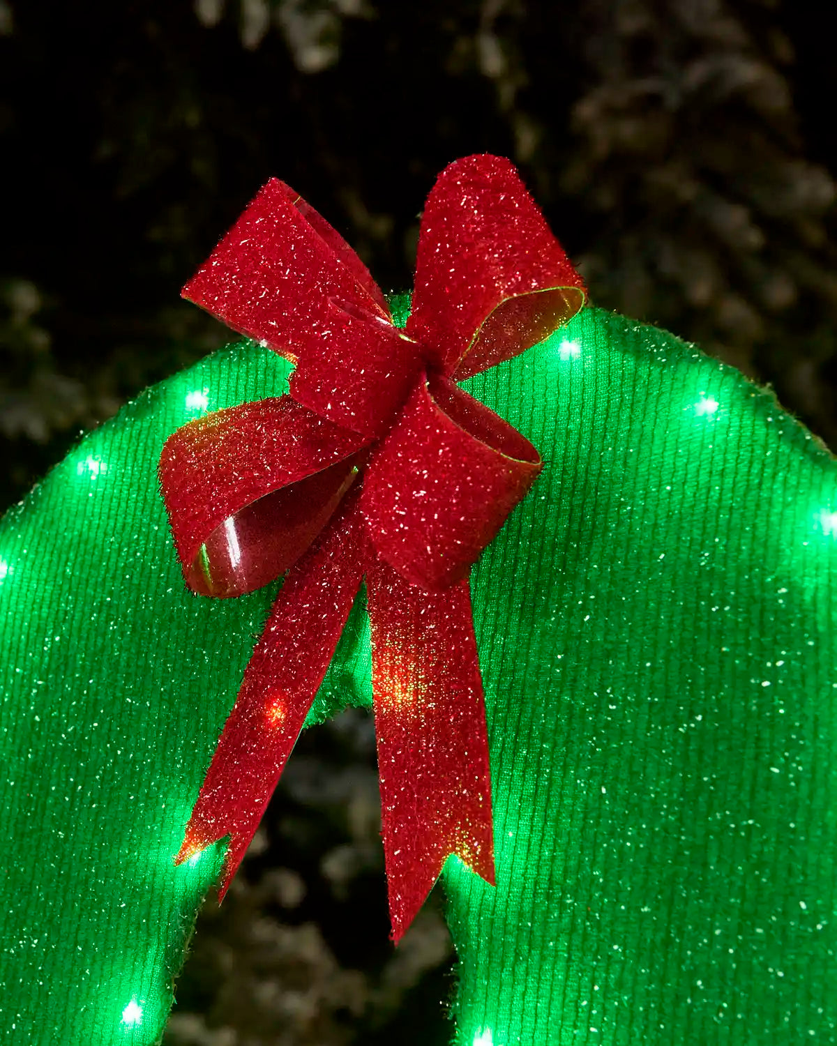 A close-up of a green glittery holiday wreath or decorative arch with a large red glitter bow on top, lit with small green lights against a dark outdoor background.