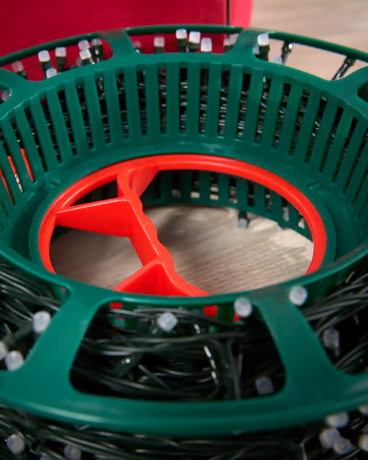 A close-up of a green plastic Christmas string light spool with a red center hub, filled with black wire and small white bulbs.