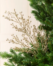 A close-up of a holiday tree branch arrangement with delicate gold glittery stems and small round gold buds, layered over green pine needles against a soft cream background.