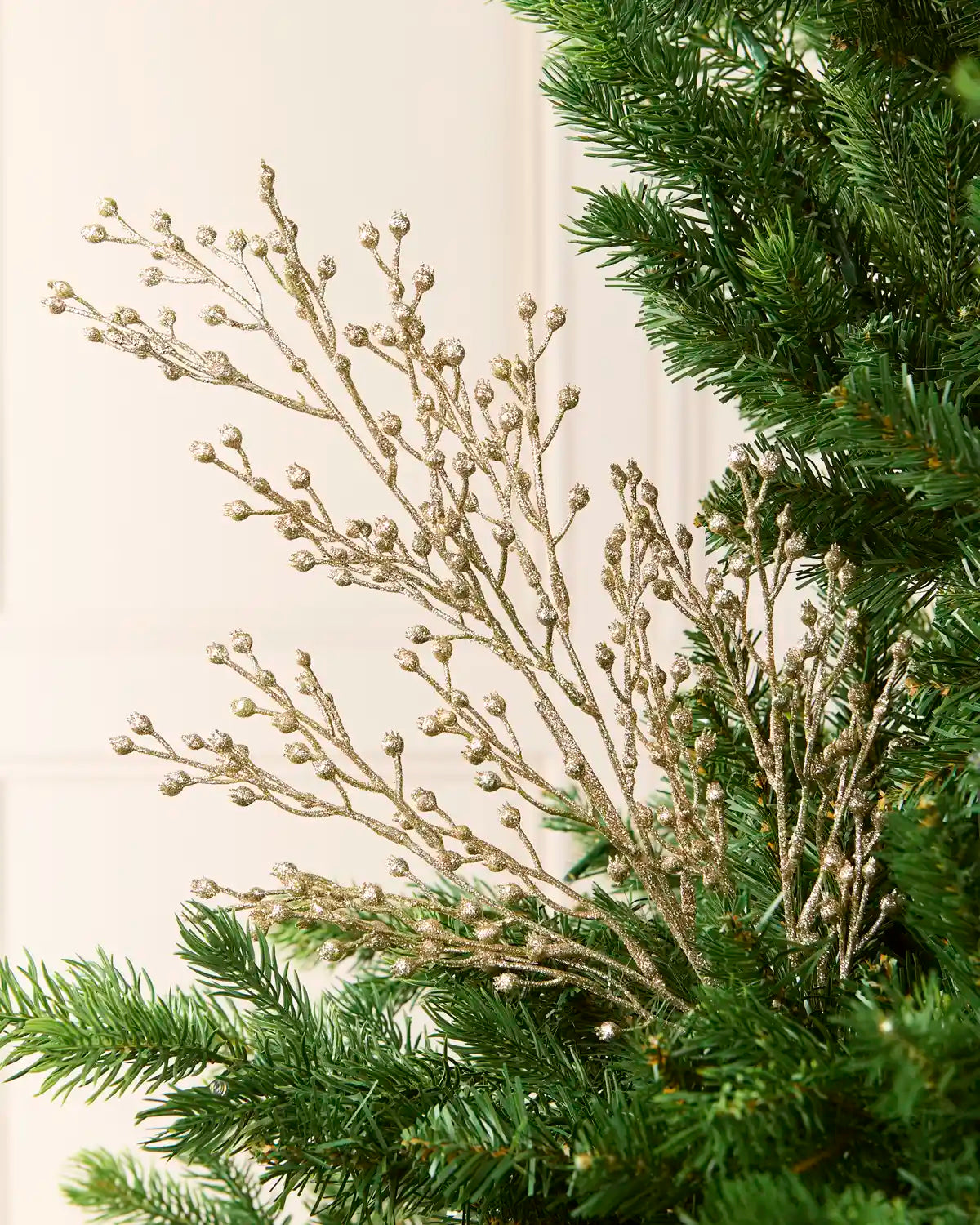 A close-up of a holiday tree branch arrangement with delicate gold glittery stems and small round gold buds, layered over green pine needles against a soft cream background.