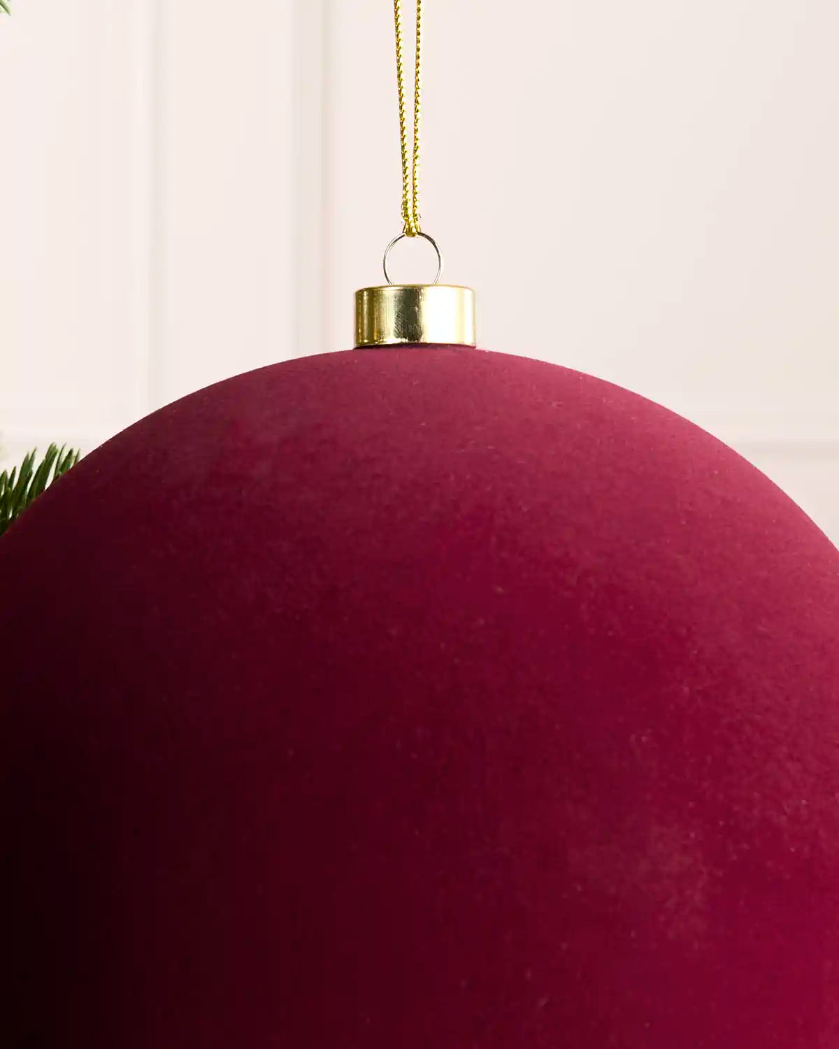 A close-up of a large, deep burgundy velvet Christmas bauble hanging from a gold cap and gold string against a soft, light background.