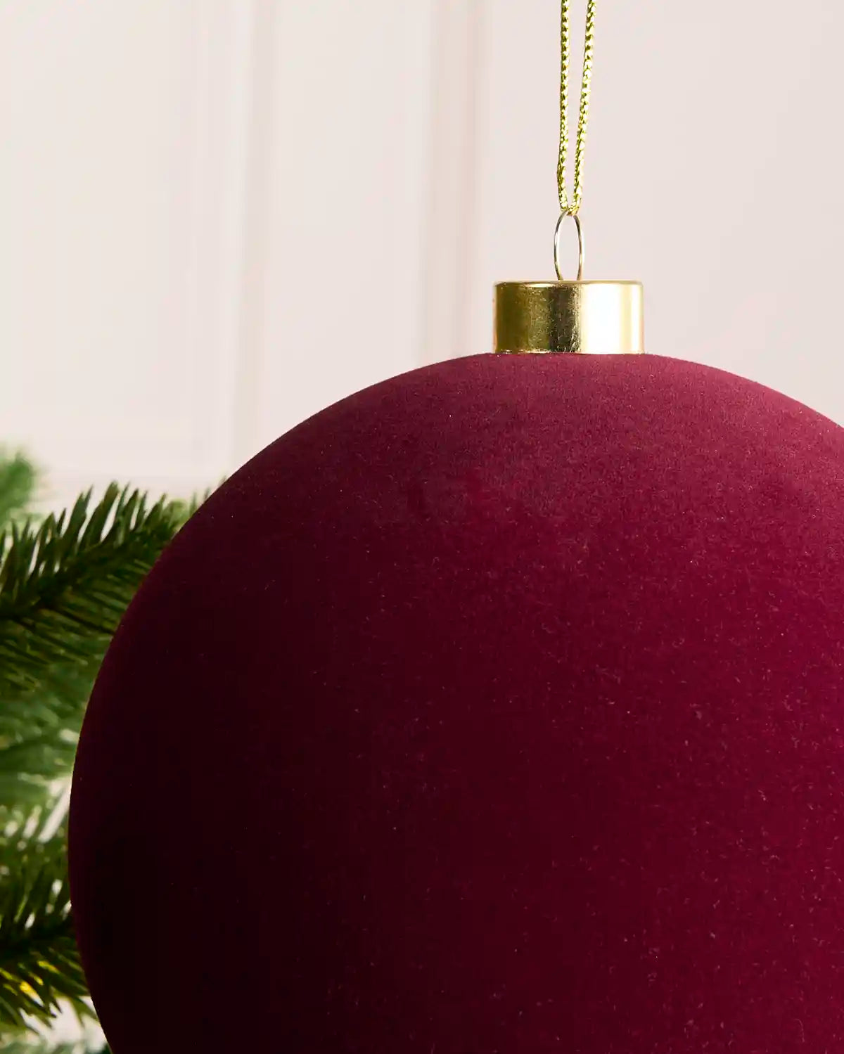 A close-up of a large, deep burgundy velvet Christmas bauble ornament with a gold cap and gold hanging string, shown next to green pine needles.
