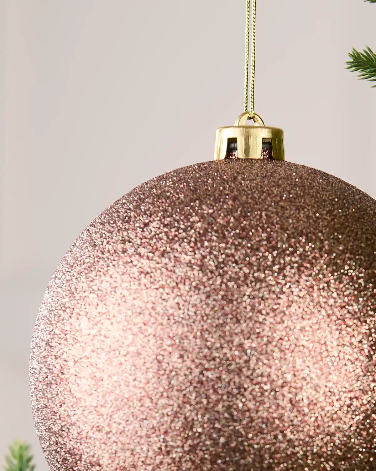 A close-up of a large glittery Mocha Christmas ornament hanging from a gold cap with a gold string, against a soft light Cream background with small green pine branches at the edges.