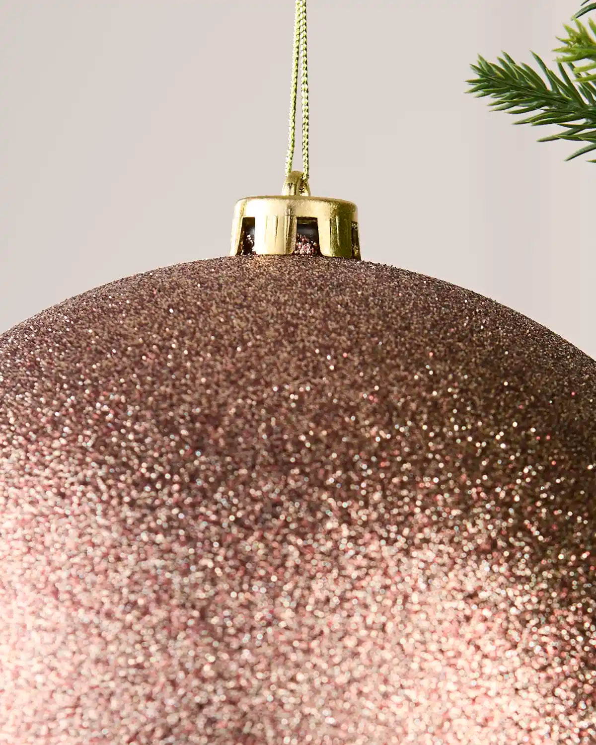 A close-up of a large glittery Mocha Christmas ornament with a gold cap and hanging string, shown against a light neutral background with a pine branch in the corner.
