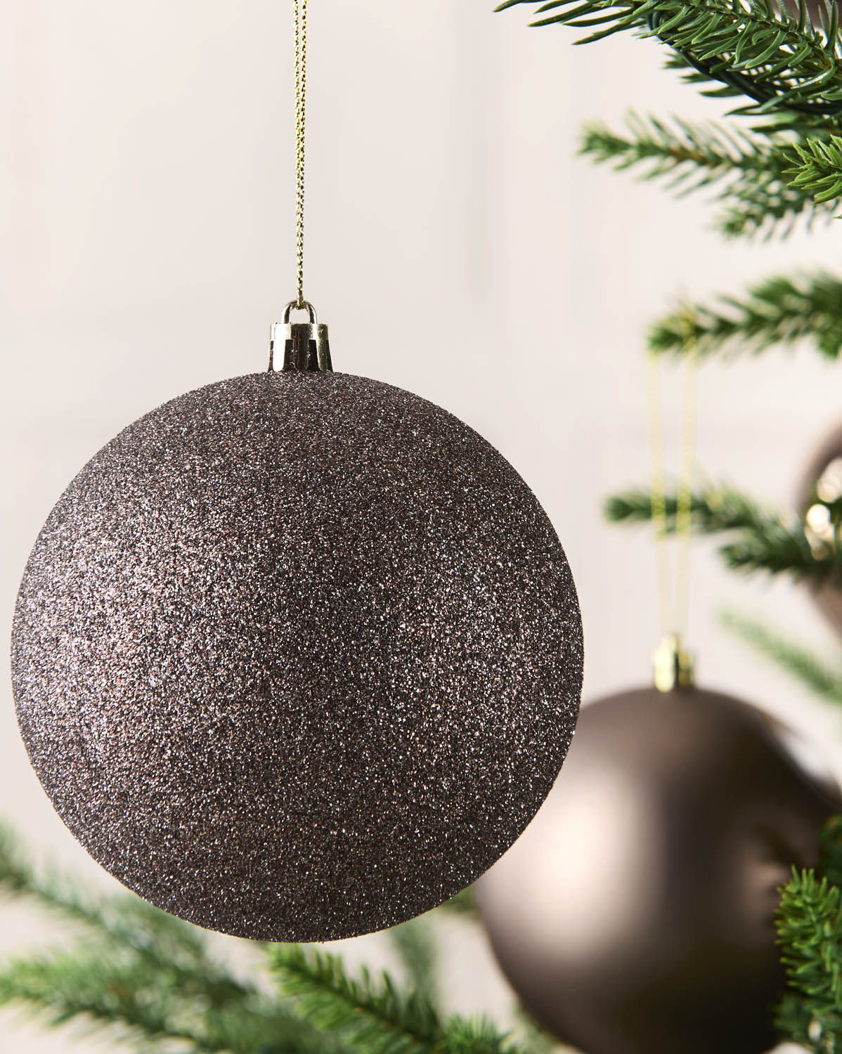 A close-up of a large, glittery Mocha Christmas tree ornament hanging from a gold string, with pine tree branches and another blurred ornament in the background.