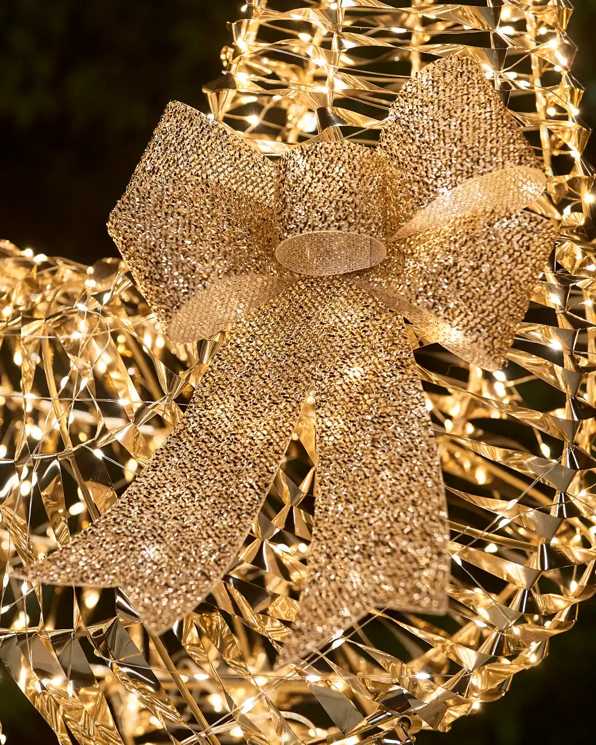 A close-up of a large glittery gold bow attached to a wire decorative holiday light structure with warm white string lights, set against a dark blurred background.
