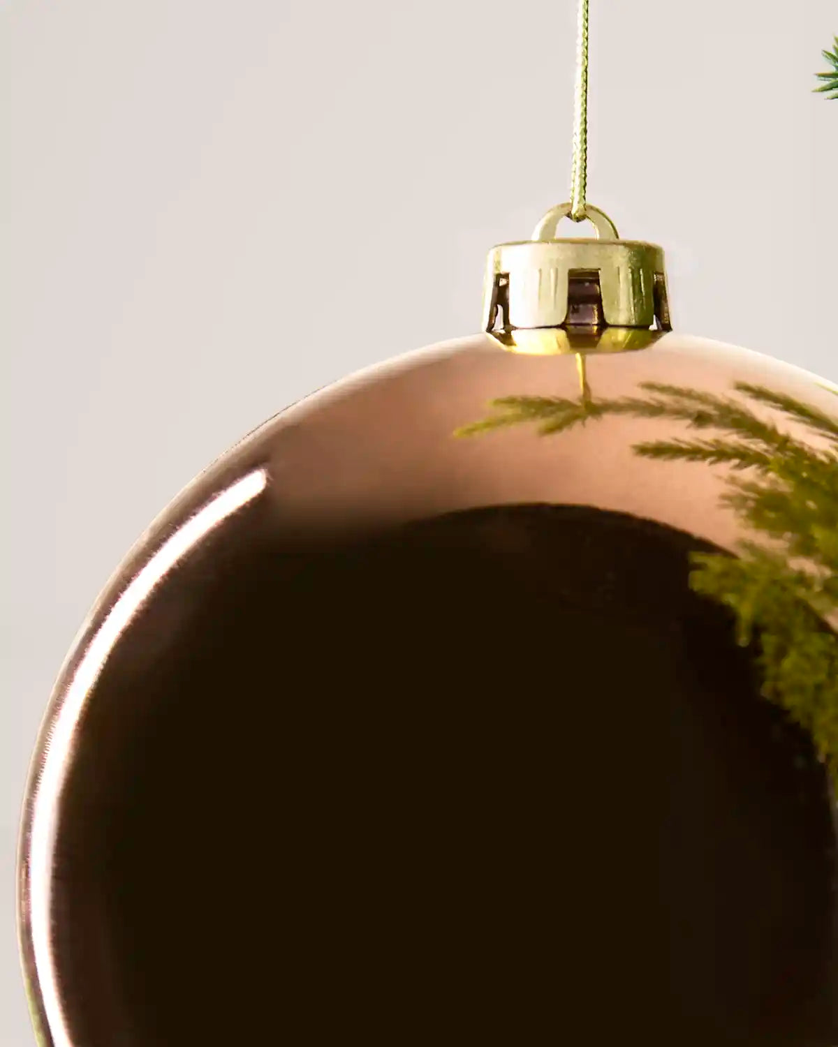 A close-up of a large, glossy Mocha-coloured Christmas bauble ornament with a gold cap and hanging string, reflecting green pine needles against a light grey background.