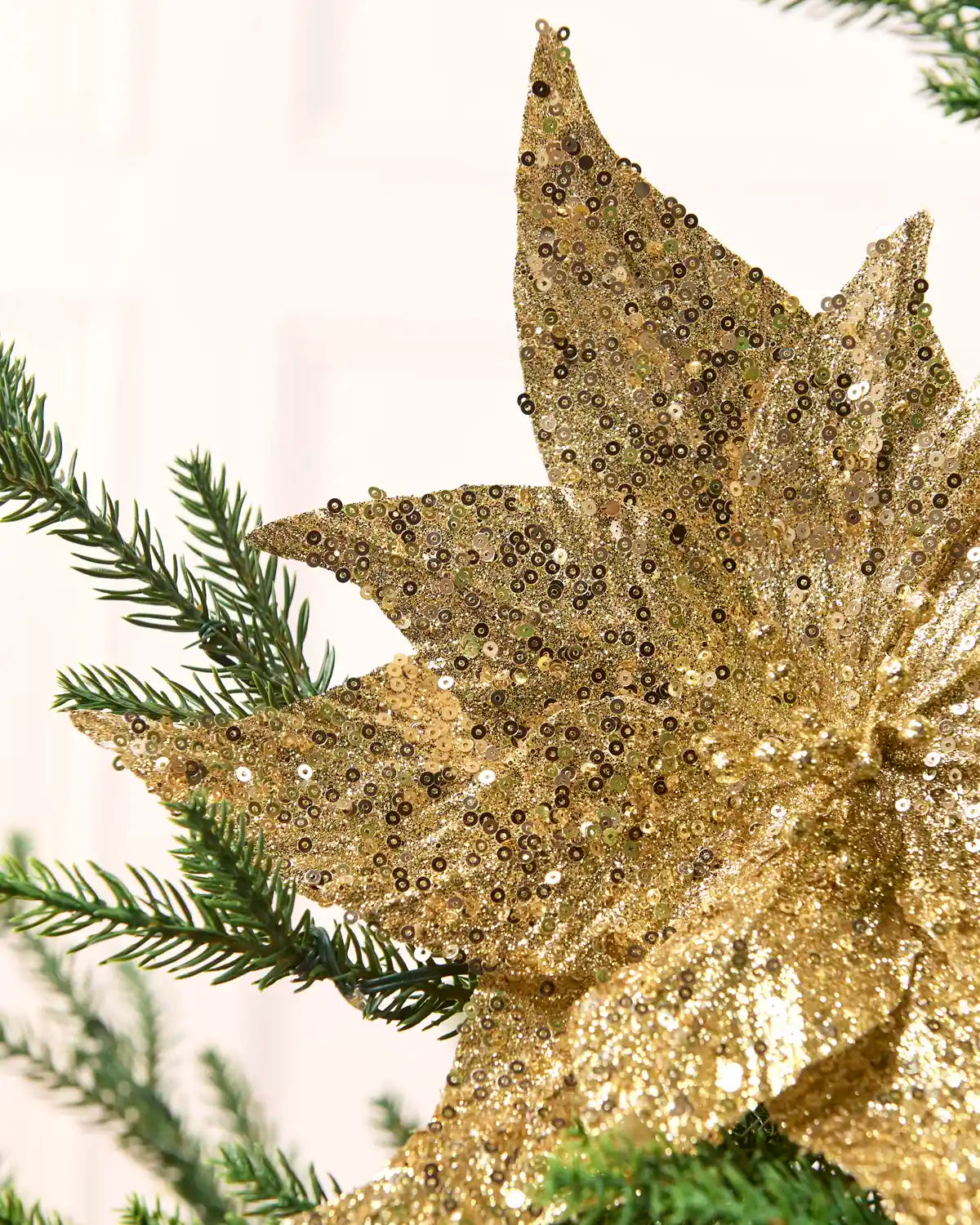 A close-up of a large gold sequined poinsettia ornament displayed among green pine branches against a soft off-white background.