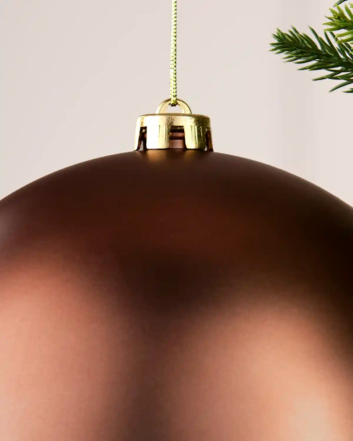 A close-up of a large, matte Espresso Christmas ornament with a gold hanging cap and string, shown against a light neutral background with a pine branch in the corner.