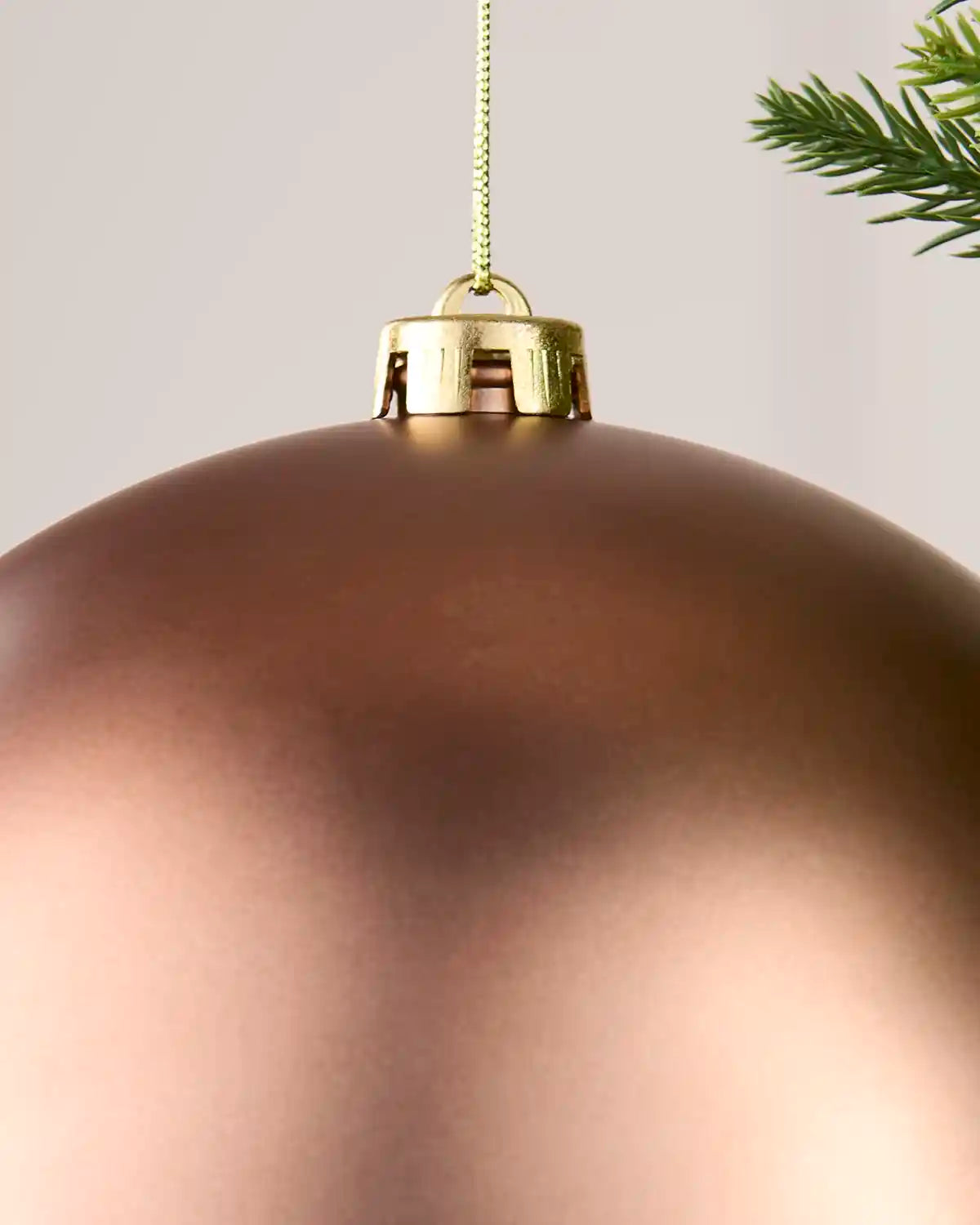 A close-up of a large, matte Mocha Christmas ornament hanging from a gold metal cap with a gold hanging string, set against a neutral light gray background with a pine branch visible in the top right corner.