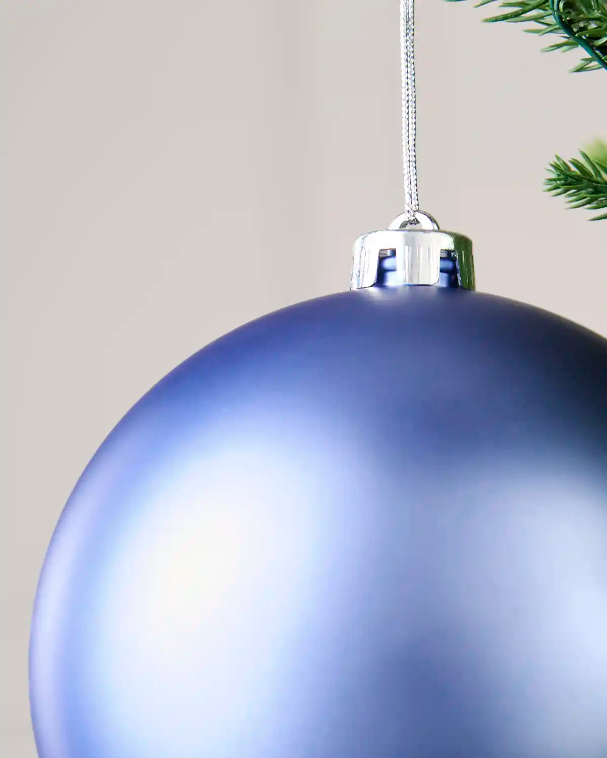 A close-up of a large, matte blue Christmas bauble ornament hanging from a silver cap on a silver string, with a hint of green pine needles in the top right corner against a neutral beige background.
