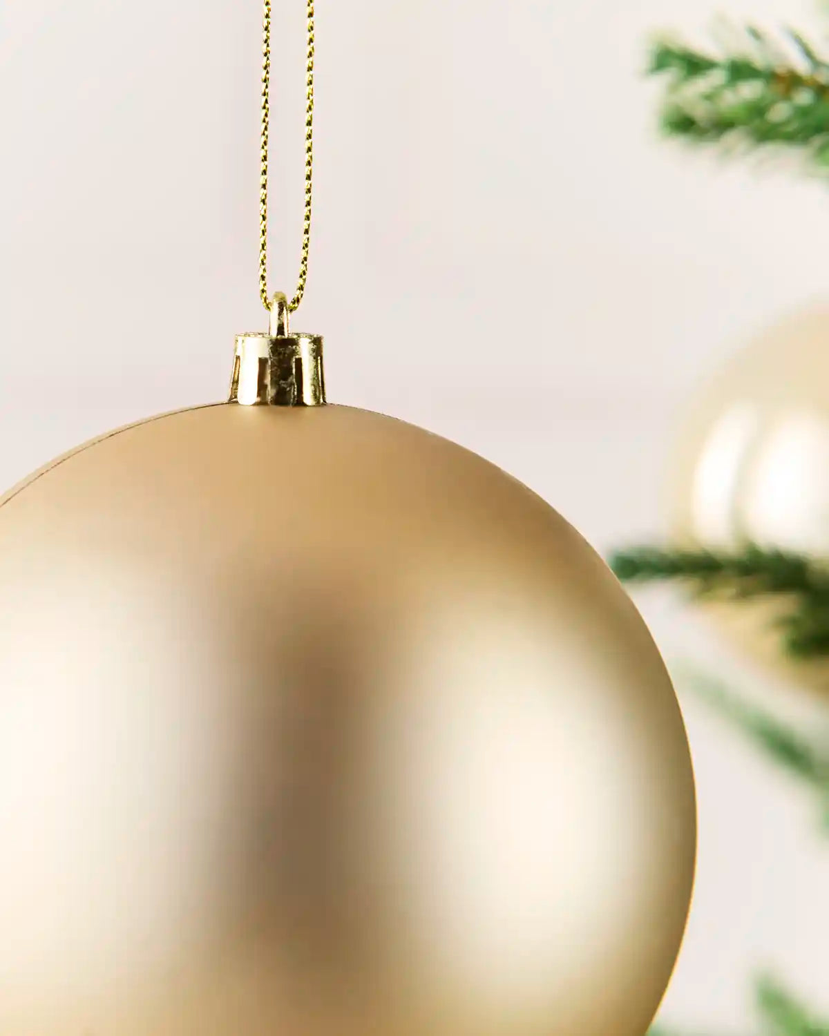 A close-up of a large, matte latte Christmas bauble hanging from a gold chain, with soft-focus green pine branches in the background.