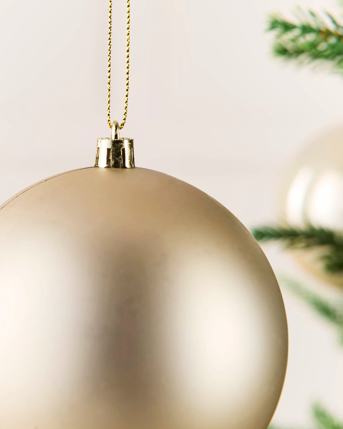 A close-up of a large, matte latte Christmas bauble hanging from a gold chain, with soft-focus green pine branches in the background.