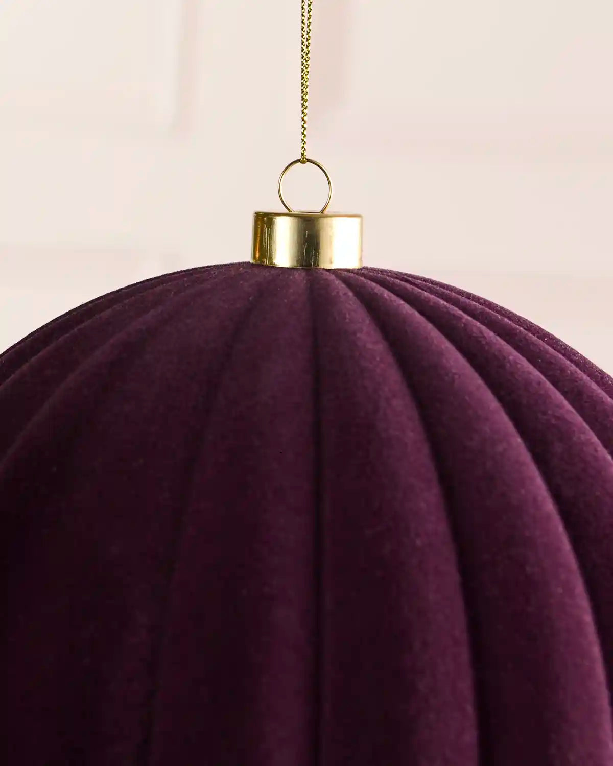 A close-up of a large pleated velvet Christmas ornament in purple, featuring a gold metal cap and a hanging gold chain against a soft cream background.