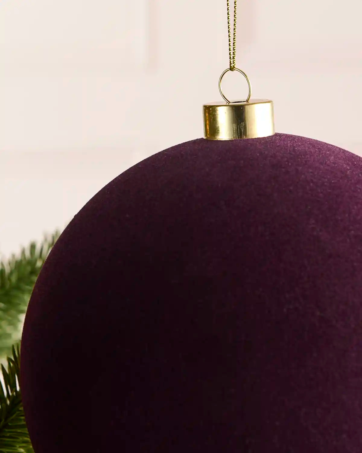 A close-up of a large, purple velvet Christmas ornament with a shiny gold metal cap and hanging loop, shown against a soft light background with a hint of green pine needles at the bottom left.