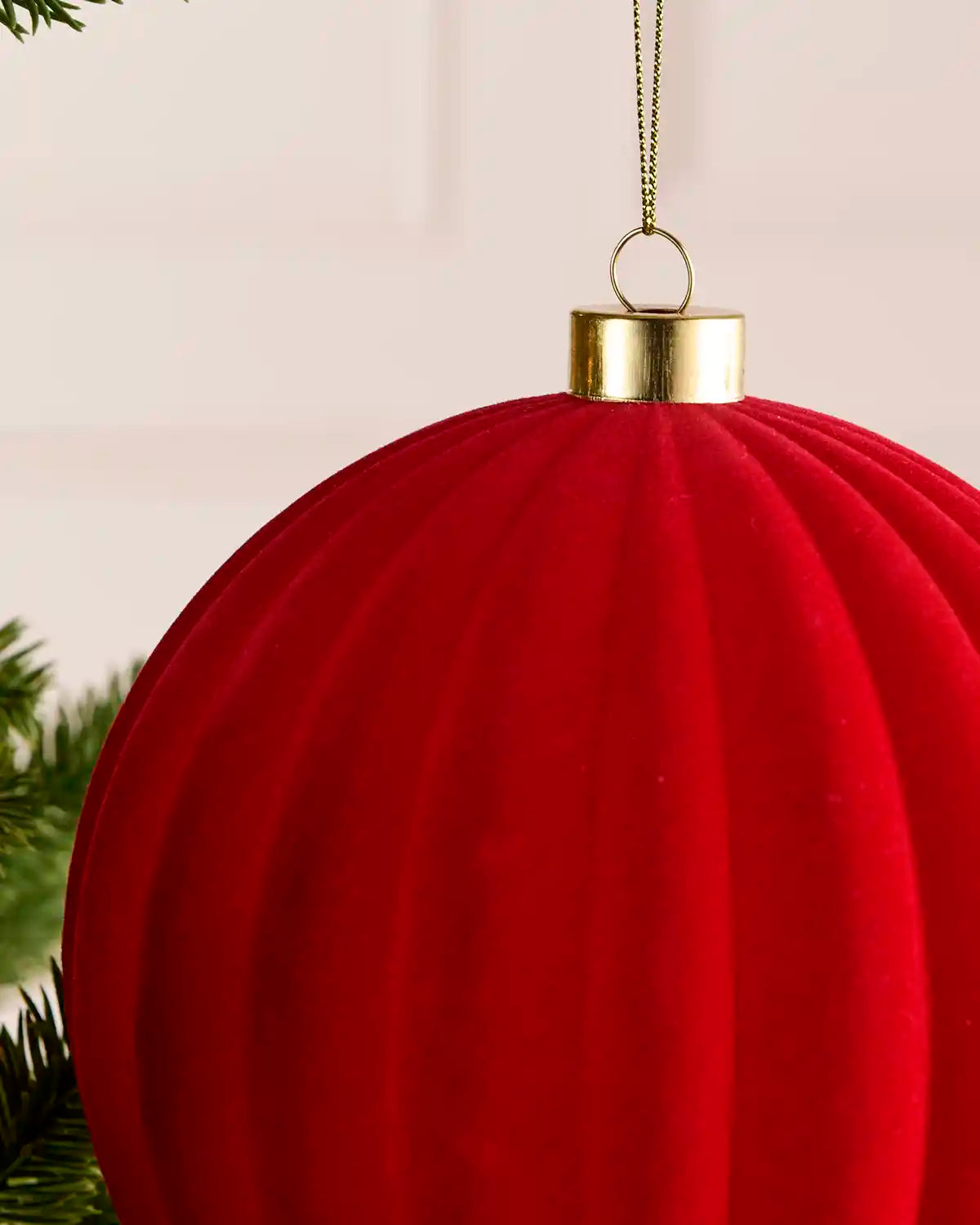 A close-up of a large red ribbed Christmas ornament hanging from a gold cap with a gold hanging loop and chain, with a soft blurred background and green pine needles at the edge.
