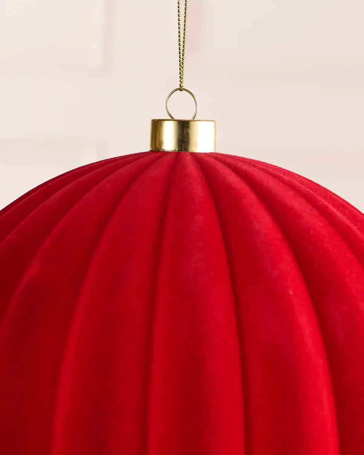 A close-up of a large red ribbed fabric ornament or hanging decoration with a gold cap and a thin gold hanging string against a soft light background.