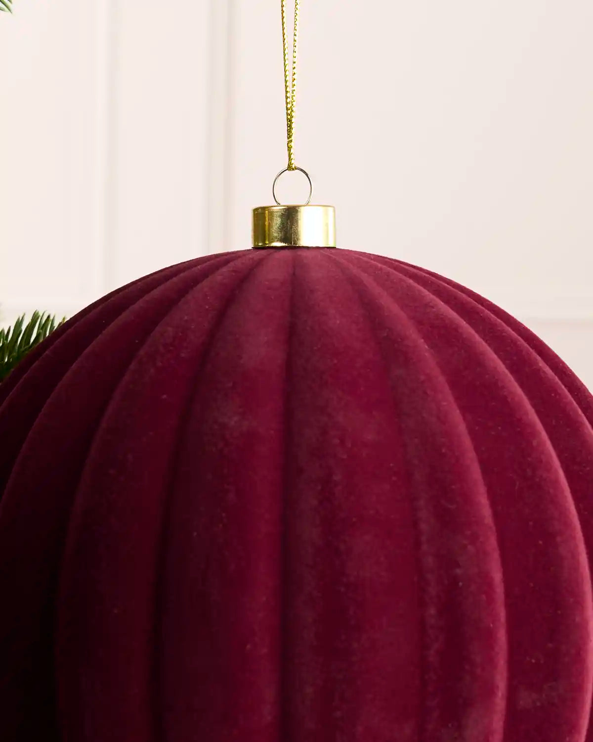 A close-up of a large ribbed velvet Christmas ornament in burgundy, hanging from a gold cap with a gold hanging string against a soft off-white background.