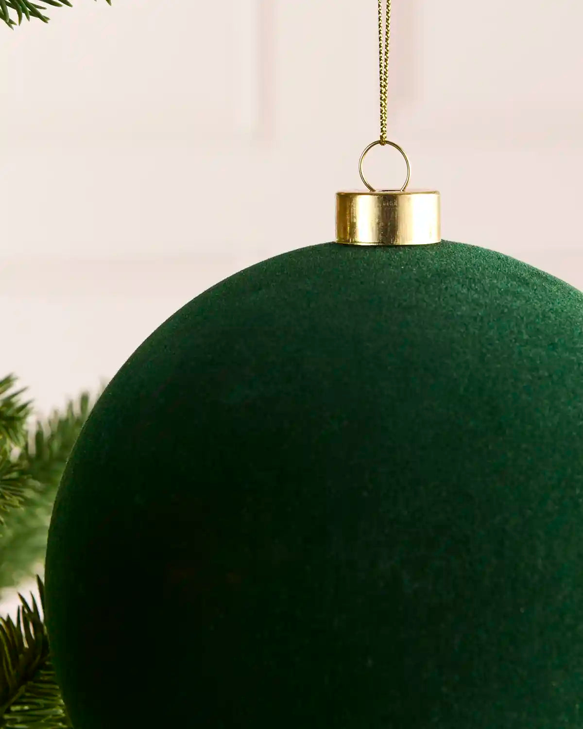 A close-up of a large sage green velvet Christmas ornament hanging from a gold cap and chain, with pine branches visible at the lower left against a soft light background.
