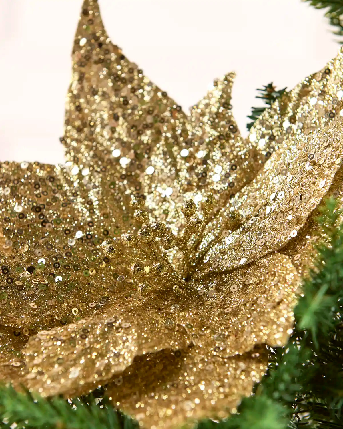 A close-up of a large, sequin gold artificial flower decoration with sparkling sequins, nestled among green pine branches against a soft light background.