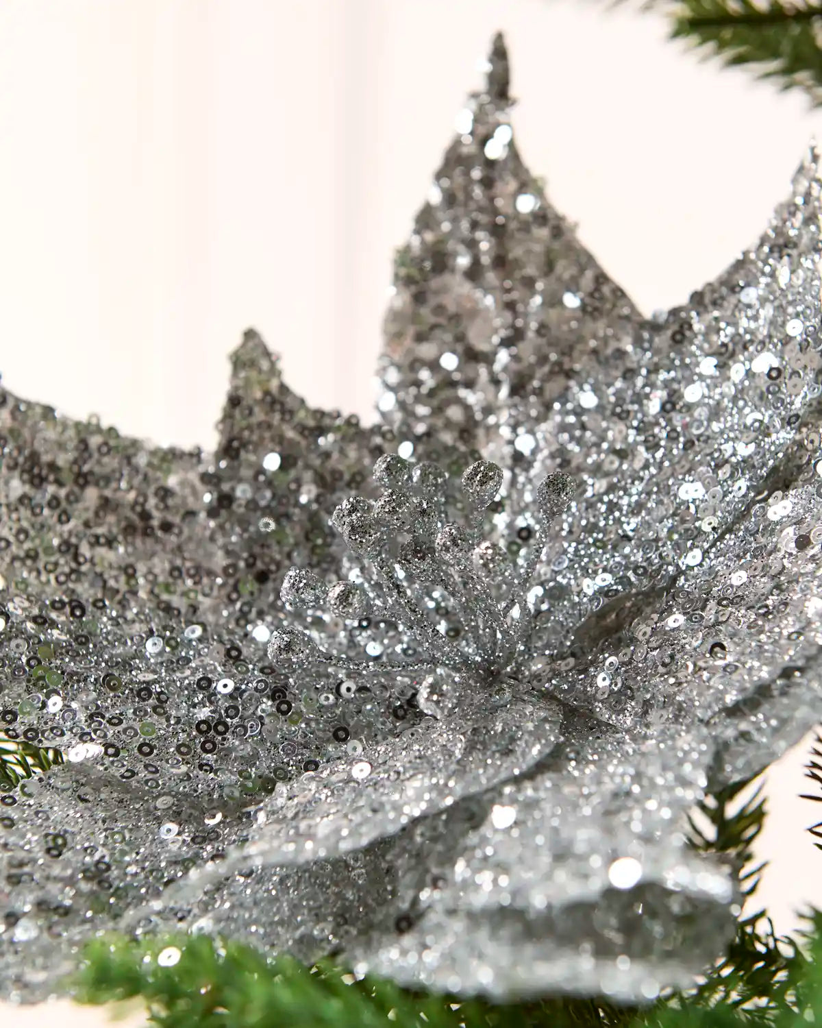 A close-up of a large silver glitter poinsettia decoration on a Christmas tree, with green pine needles visible at the bottom and background.