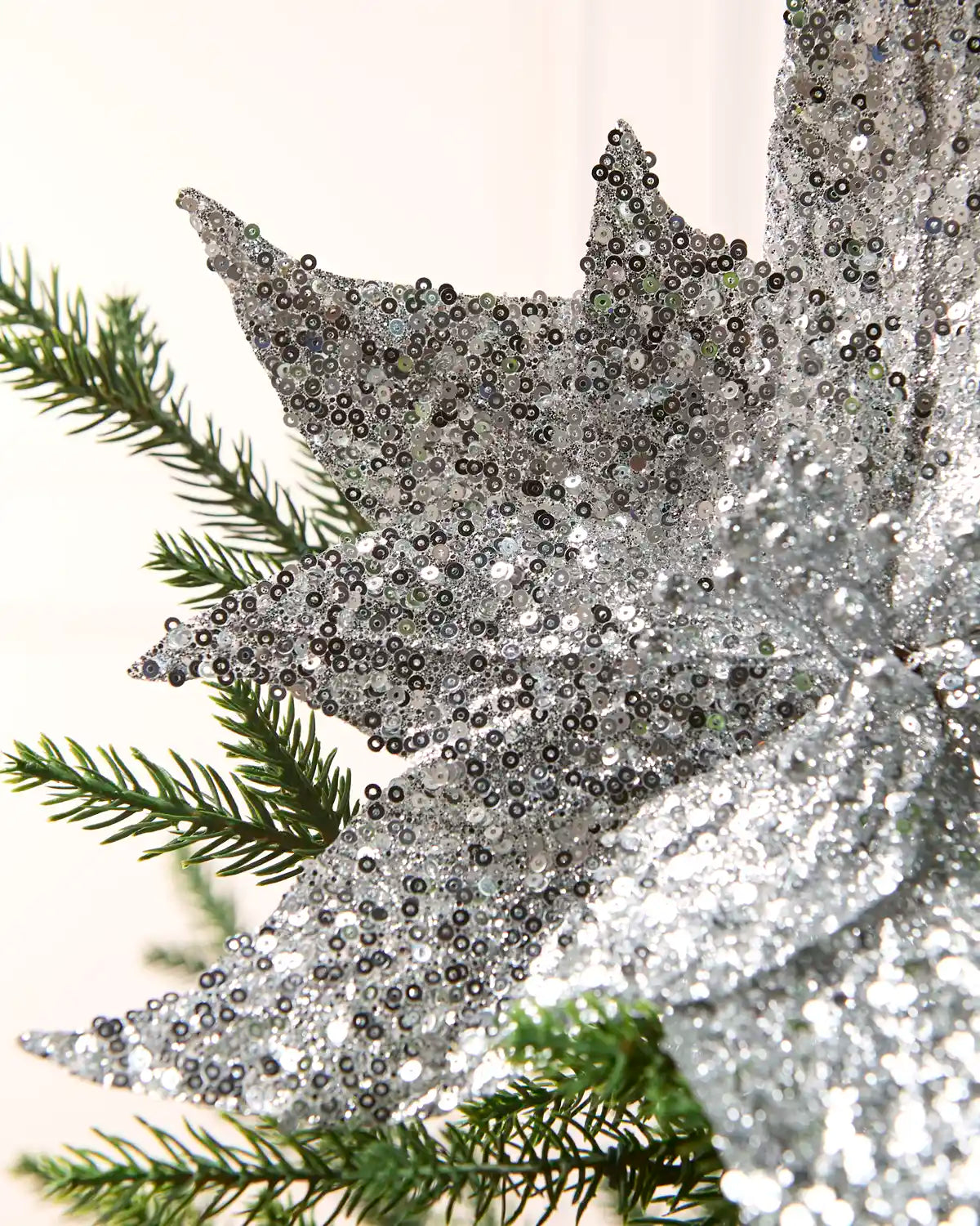 A close-up of a large silver sequined decorative poinsettia-style ornament with green pine branches layered behind it against a soft off-white background.