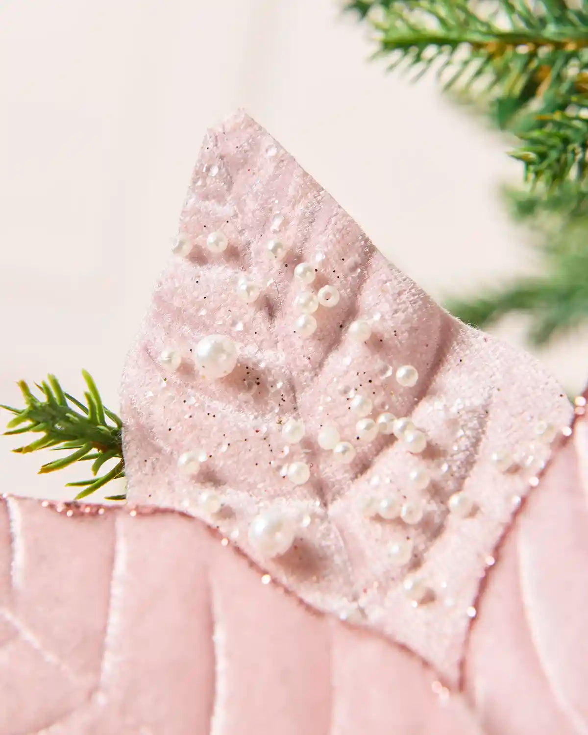 A close-up of a light pink velvet leaf on a poinsettia, adorned with white faux pearls and glitter, styled with green pine branches.