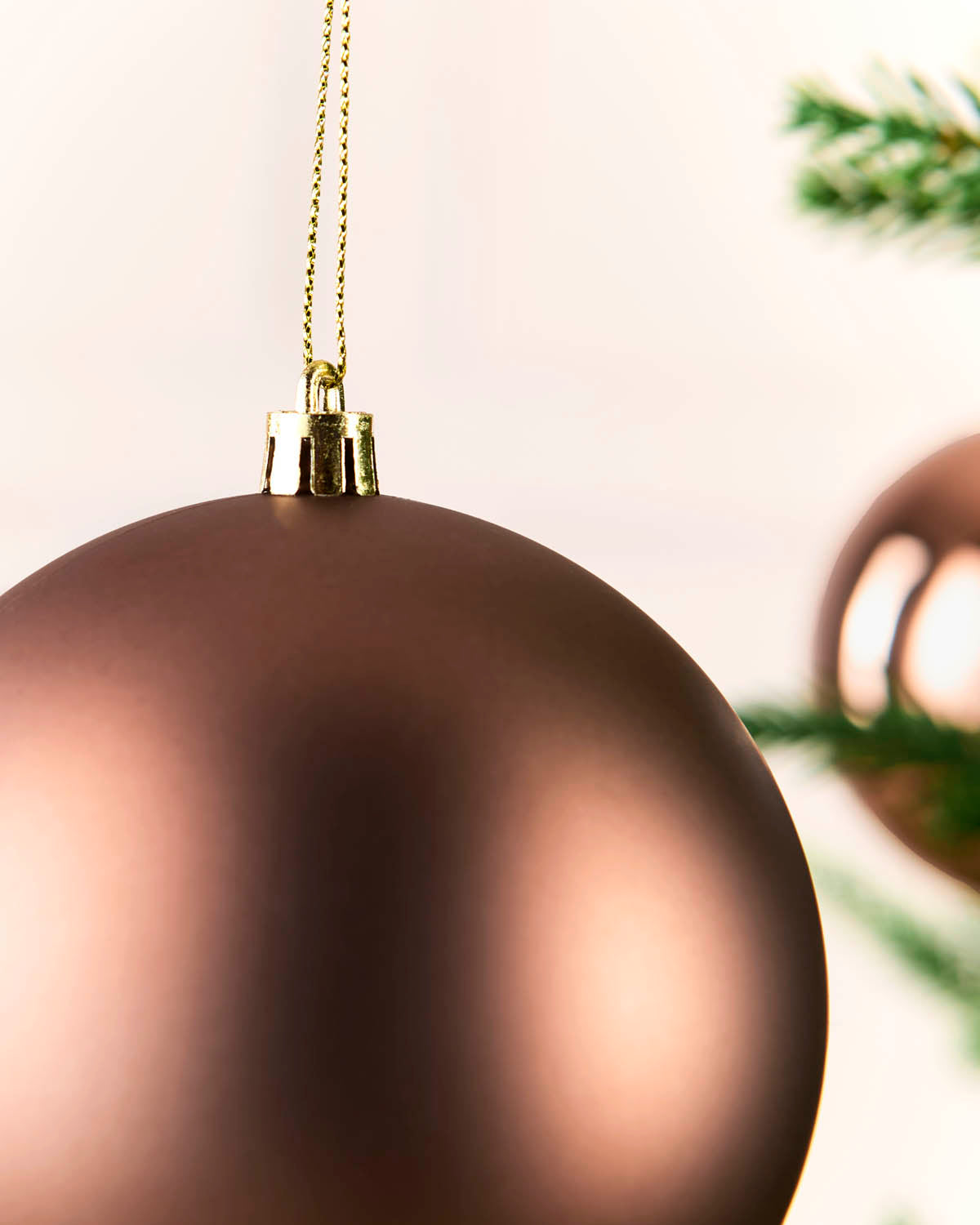 A close-up of a matte espresso Christmas ornament hanging by a gold string, with soft-focus pine branches in the background.