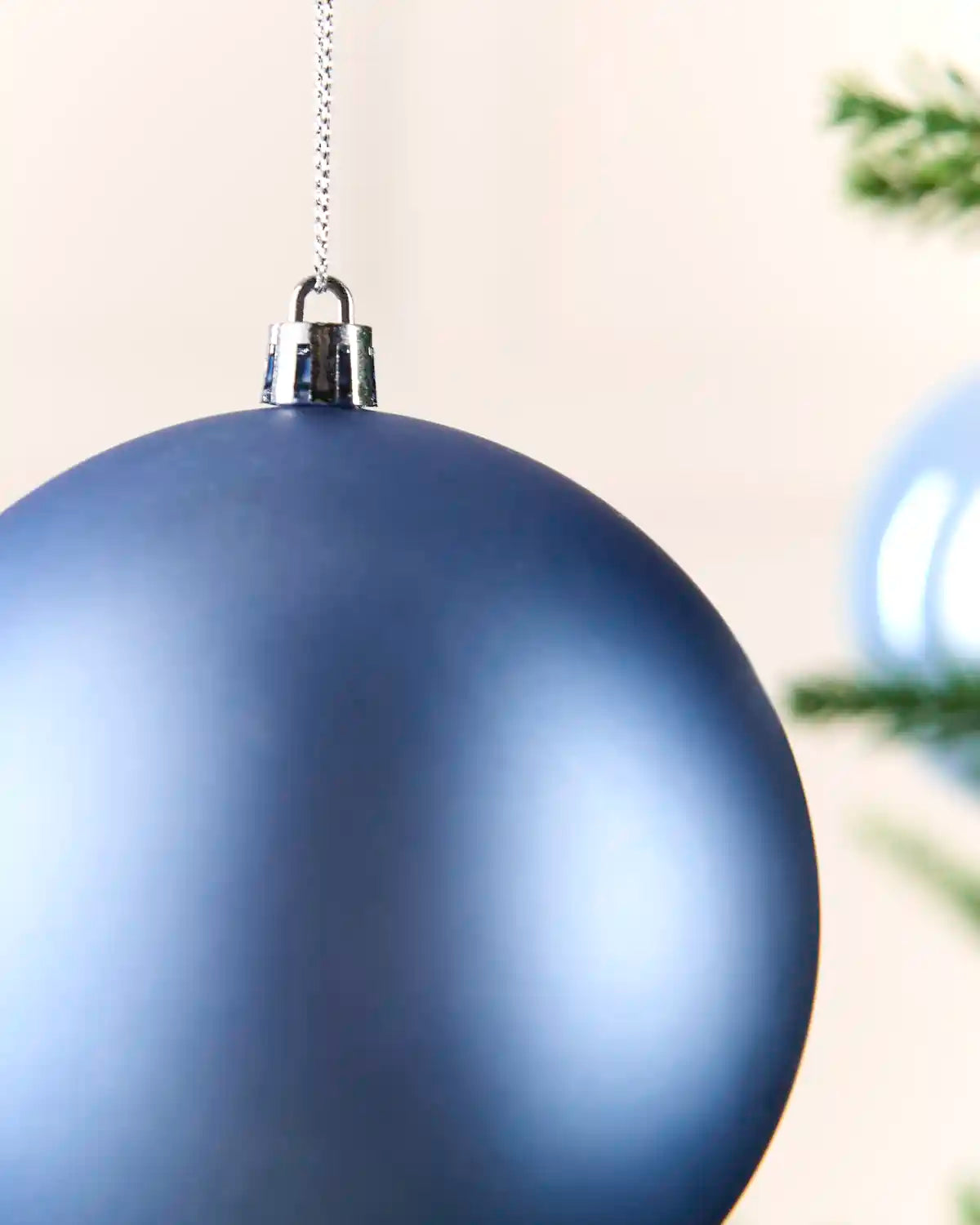 A close-up of a matte ice blue Christmas bauble ornament hanging from a silver chain against a soft beige background with blurred green pine branches.
