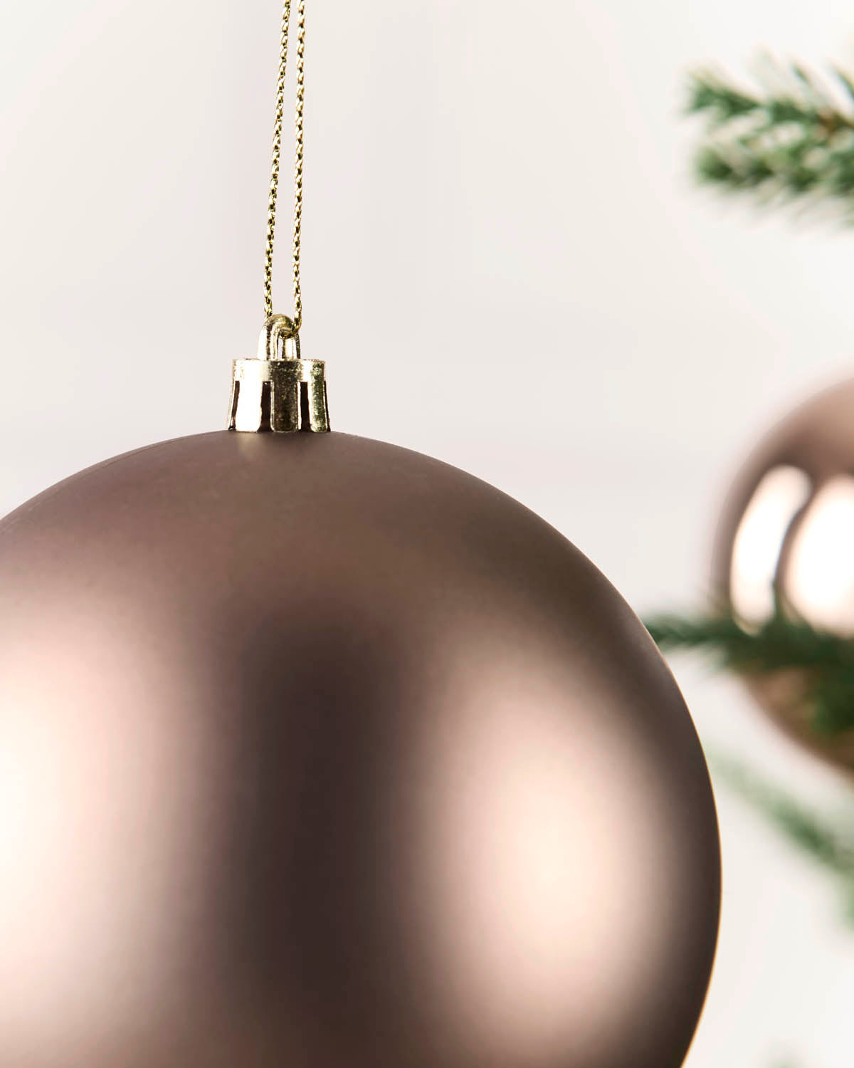A close-up of a matte metallic Mocha Christmas bauble hanging from a gold wire hanger, with a soft-focus pine branch in the background.