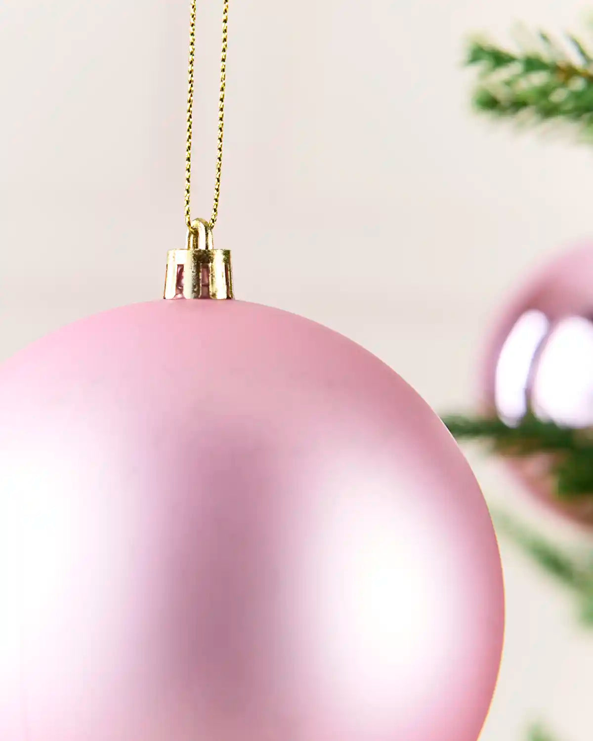 A close-up of a matte pink Christmas bauble hanging from a gold chain, with a softly blurred green pine branch in the background.