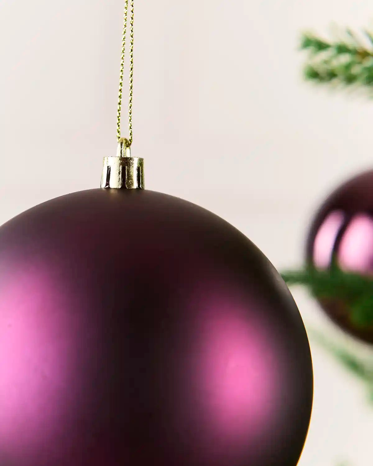 A close-up of a matte purple Christmas bauble hanging from a gold string, with a blurred second ornament and green pine branches in the background.