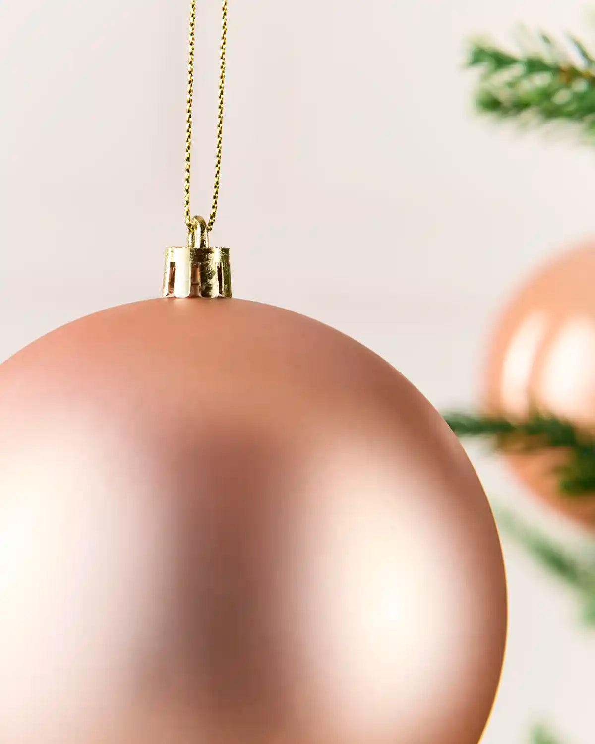 A close-up of a matte rose gold Christmas bauble hanging from a gold chain, with soft-focus pine branches in the background.