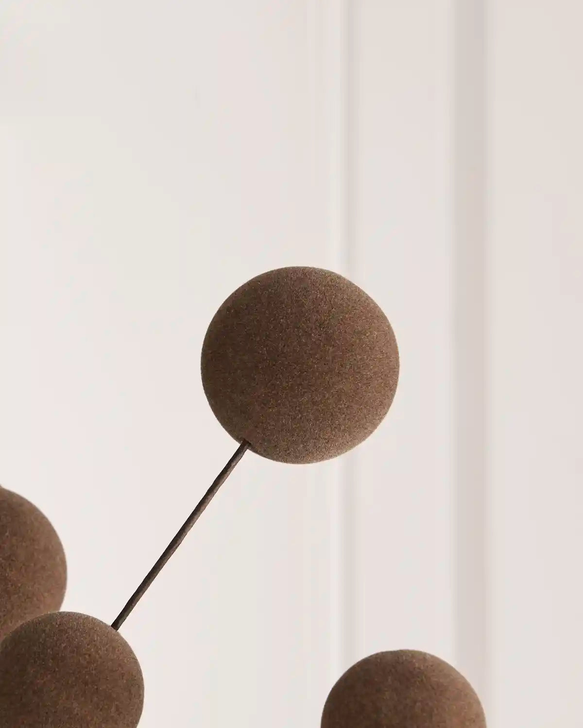 A close-up of a modern decorative mobile or wall hanging with matte brown spherical elements on thin dark metal rods against a soft off-white background.