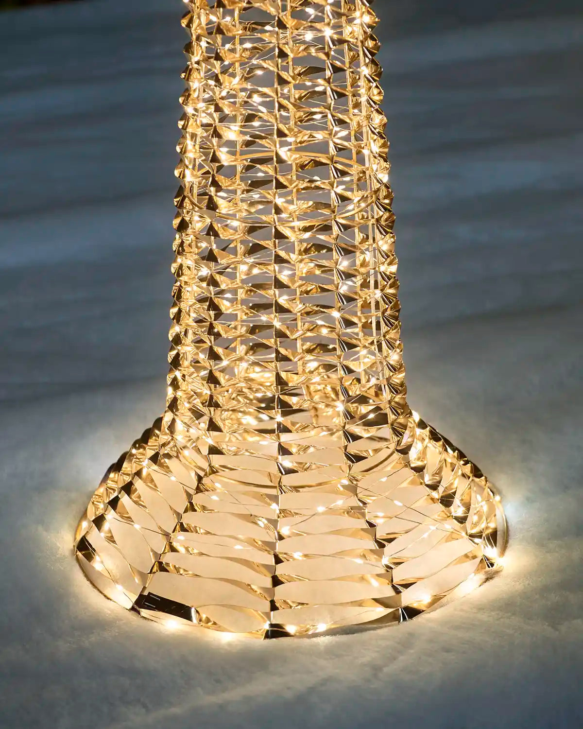 A close-up of a modern sculptural table lamp with a flared base and a tall, tapered body made of interlocking metallic rings that glow warm yellow light. The lamp is set against a dark, textured background.