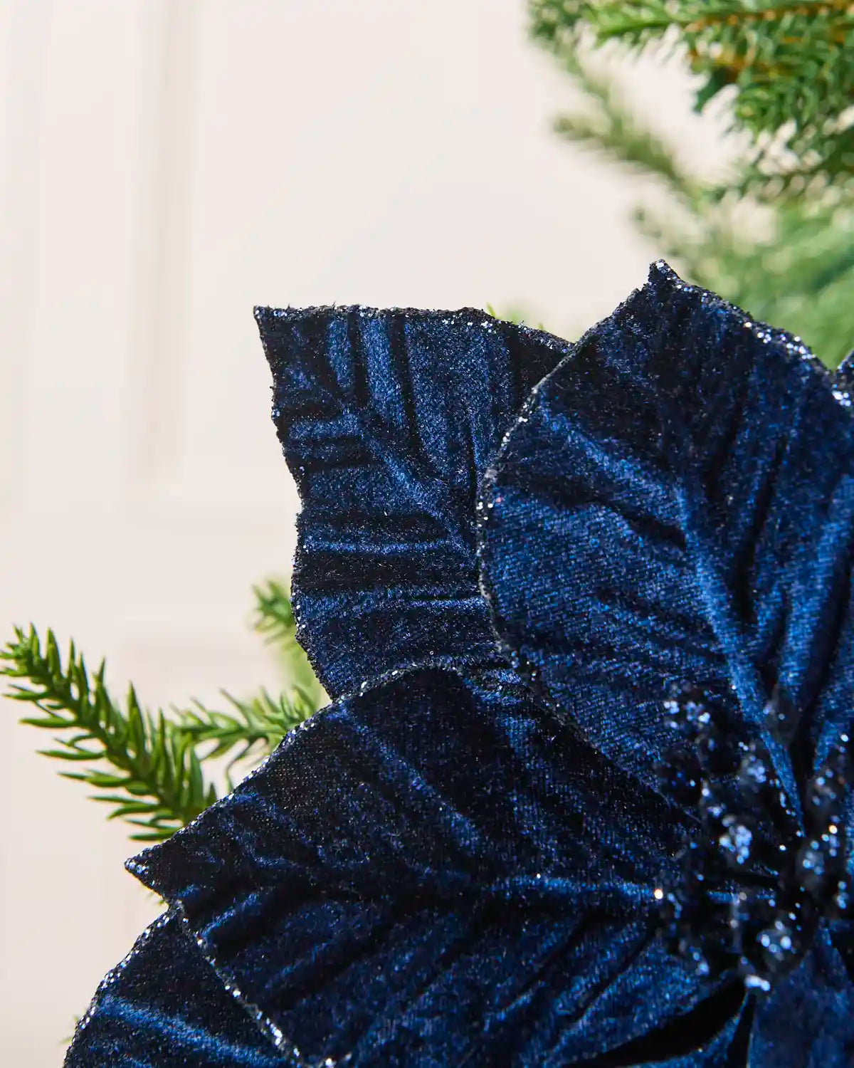 A close-up of a navy blue poinsettia flower decoration with large textured petals and small green sprigs against a cream panelled background.

