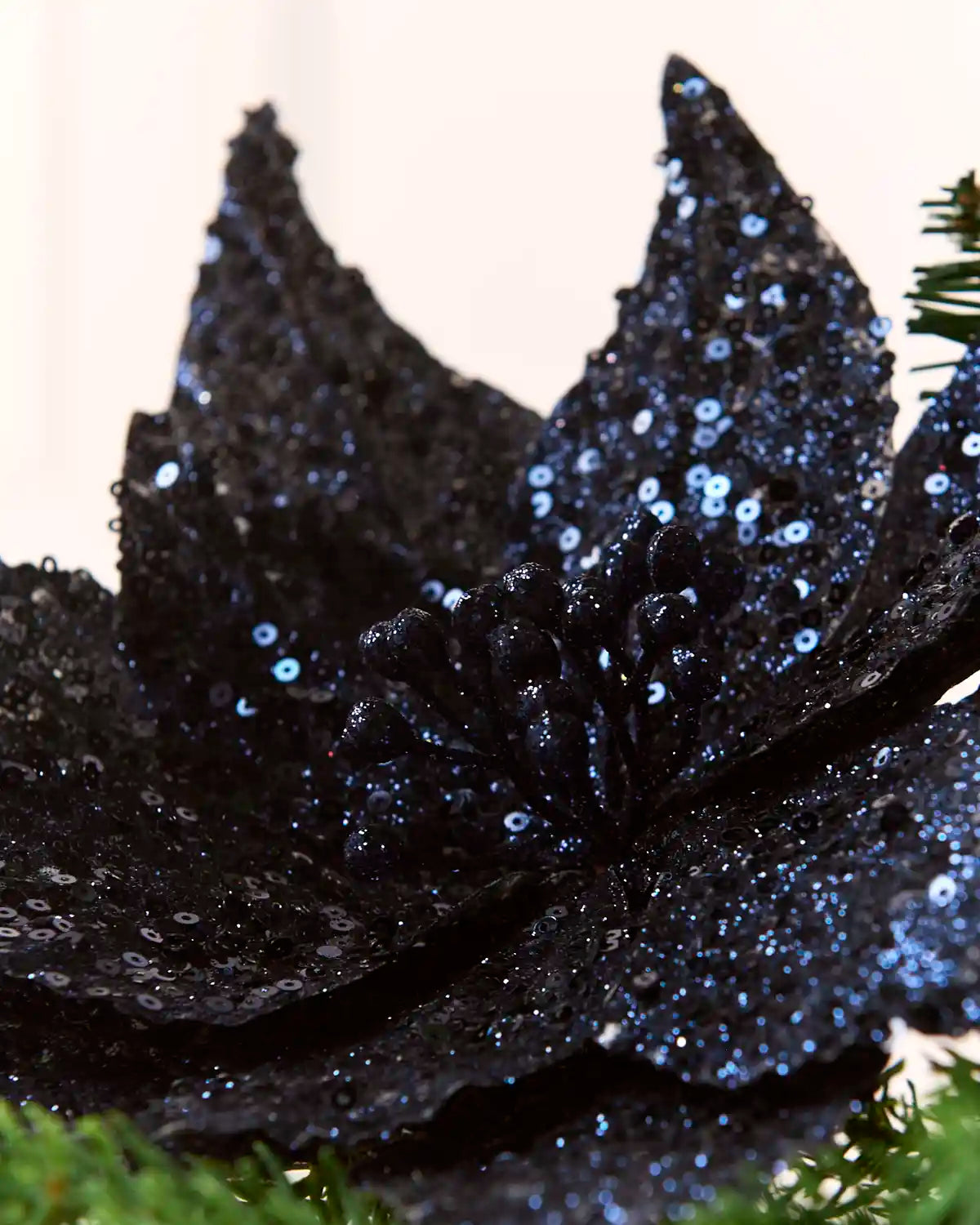 A close-up of a navy sequined decorative poinsettia  flower ornament with blue reflective sequins, set against a soft light background with green foliage at the edges.